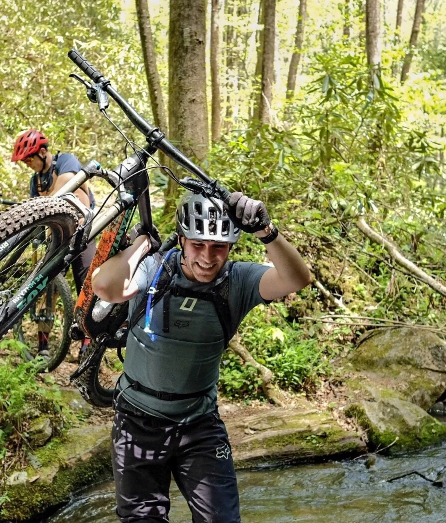 A grinning rider carries his mountain bike across a creek, another rider behind him.